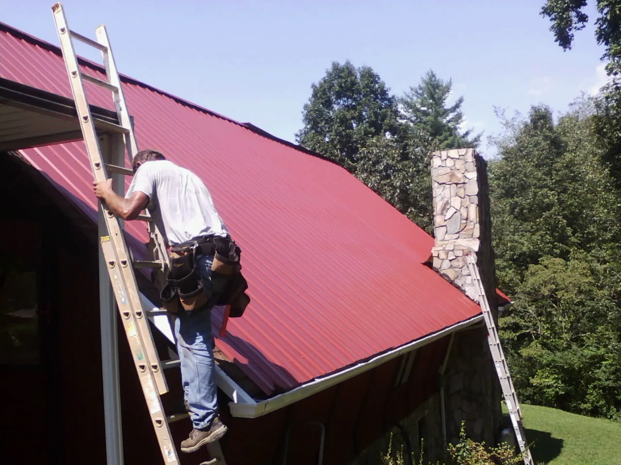 Roofer on a ladder beside a red metal roof and stone chimney — Soffit & Fascia Repair by Roofing Brothers in Forest Lakes